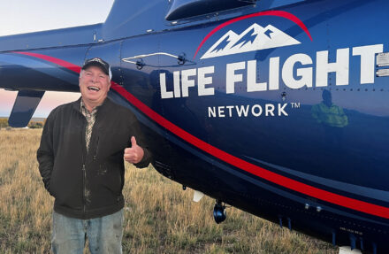 An adult stands outdoors beside the side of a blue Life Flight Network helicopter, giving a thumbs-up gesture. The helicopter’s fuselage fills most of the frame, with the “Life Flight Network” logo and mountain graphic clearly visible against a grassy field and open sky.