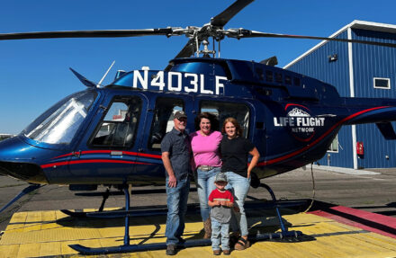 Four people pose on a helipad in front of a blue Life Flight Network helicopter with the registration number N403LF visible on the fuselage. Three adults stand side by side near the helicopter’s landing skids, while a small child stands in the foreground. The helicopter’s rotor blades, windows, and medical transport branding are visible, with a blue hangar and clear sky in the background.