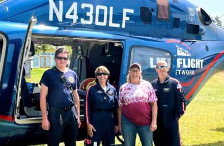 Four adults stand on a grassy area beside an open blue medical helicopter marked with “Life Flight Network” branding and registration number N430LF. The helicopter’s side door is open, showing part of the interior, with the fuselage, windows, and landing skids visible in bright daylight.